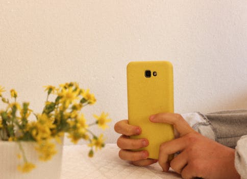 Hands holding a smartphone with a yellow case next to a vase of flowers indoors.