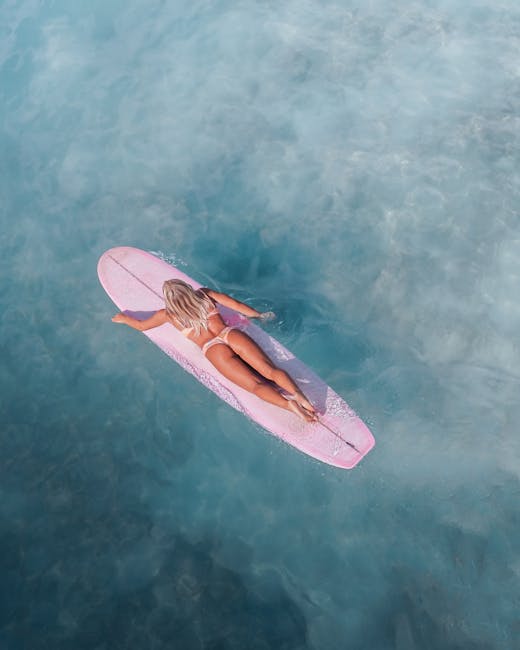 Overhead shot of a woman surfing on a pink surfboard over clear blue water, exuding a sense of adventure and freedom.