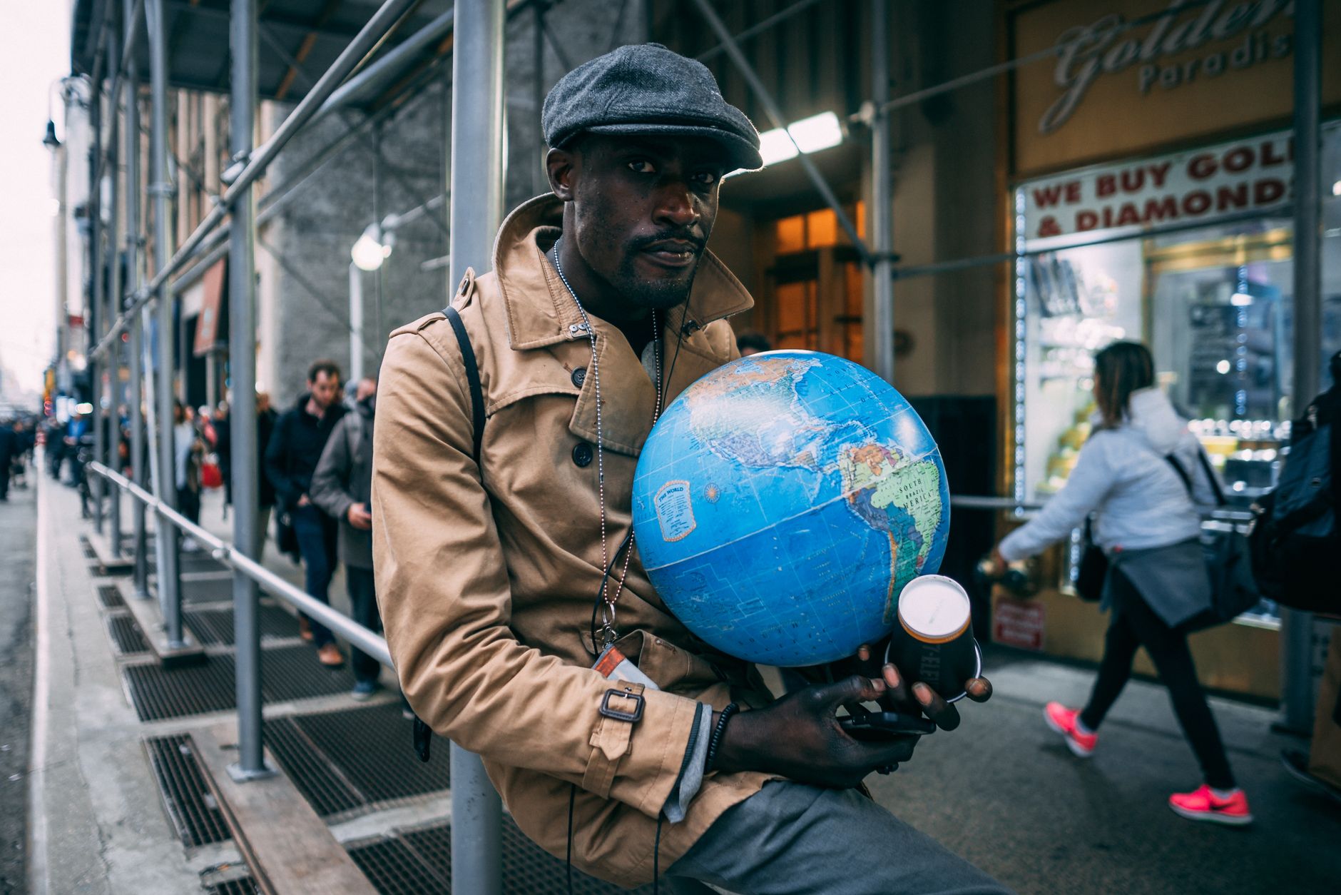 Man in flat cap and coat holding a globe on a bustling city sidewalk.