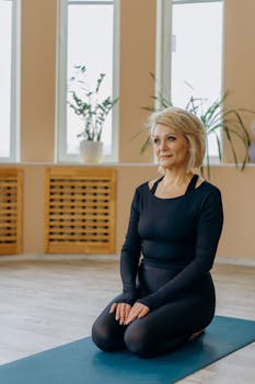 Senior woman in activewear kneels on yoga mat in bright room, embodying fitness and lifestyle.