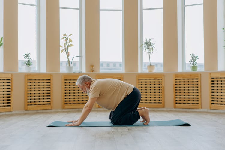 An Elderly Man In Beige T-shirt And Black Pants Kneeling On Floor With Yoga Mat