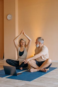 Elderly couple practicing yoga together indoors, following an online tutorial on a laptop.