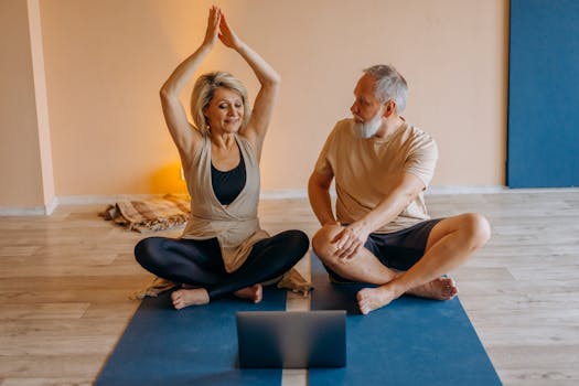 Senior couple practicing yoga together on mats indoors, using a laptop for guidance.