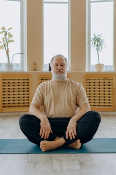 Elderly man sitting cross-legged on yoga mat indoors, meditating and relaxing.