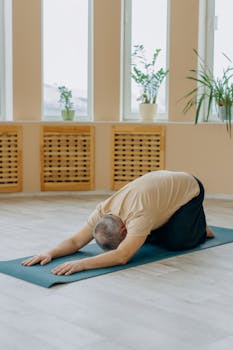Elderly man performing a forward bend yoga pose on a mat in a bright indoor space.