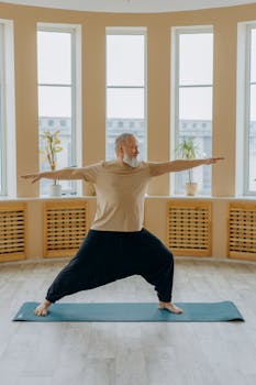 Senior man doing yoga on a mat with arms extended in a sunlit room. Promotes healthy lifestyle and fitness.