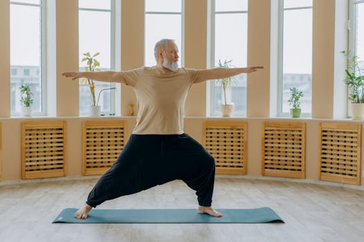 Elderly man with white hair doing yoga on mat inside bright room.