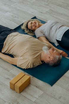 Elderly couple lying on yoga mats, smiling and enjoying relaxation indoors.