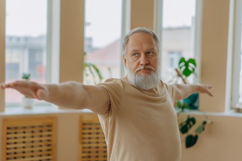 Free Elderly man performing Warrior Pose yoga for wellness indoors. Stock Photo