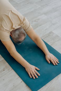A senior man practicing a forward bend yoga pose on a blue mat indoors, promoting fitness.