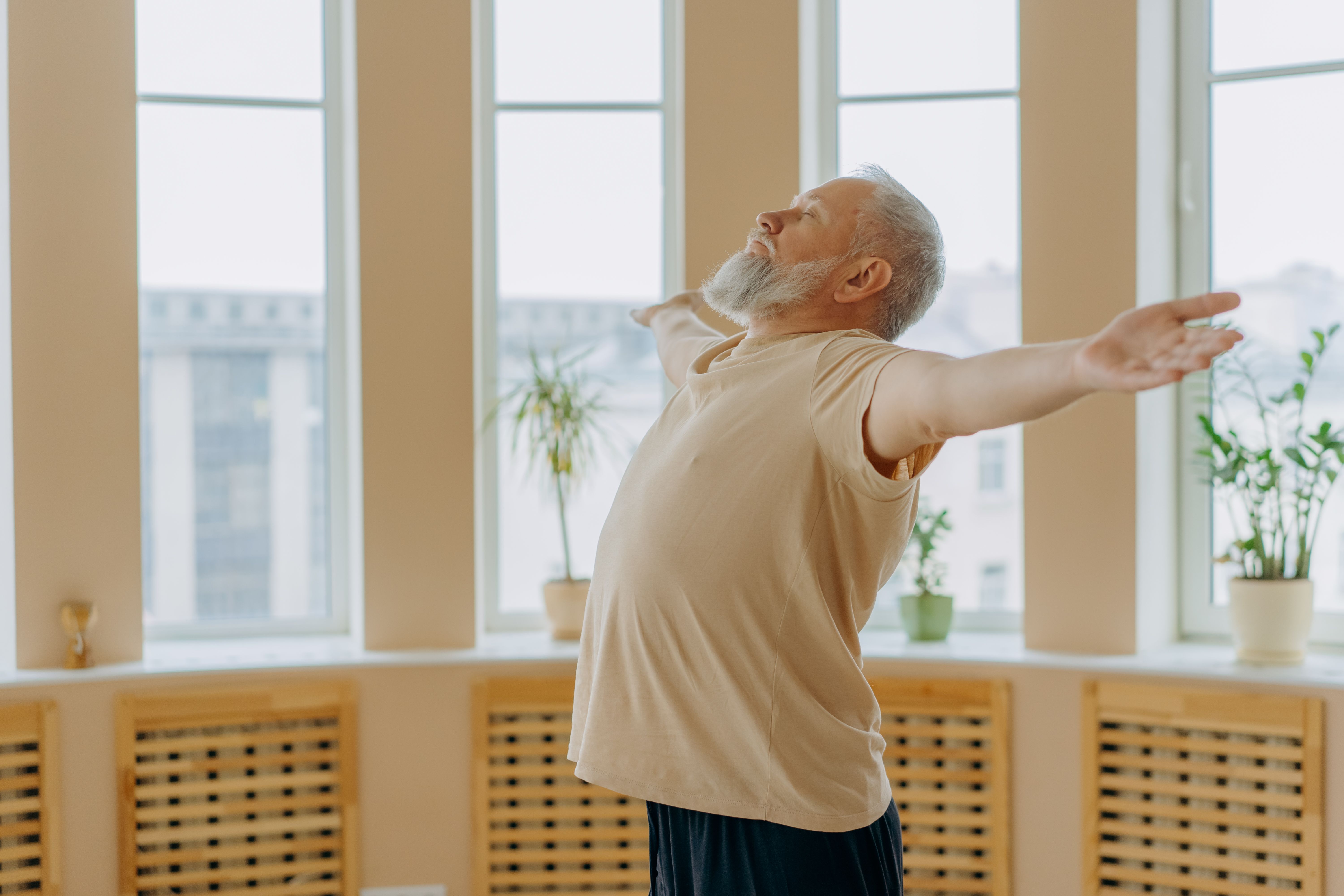 Elderly man meditating with eyes closed, arms wide in an indoor space, expressing calm.