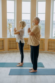An elderly couple stands on yoga mats performing a pose in a bright room.