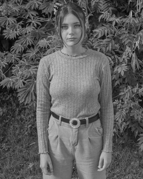 Grayscale Photo of Young Woman in Long Sleeve Sweater Standing Near Trees