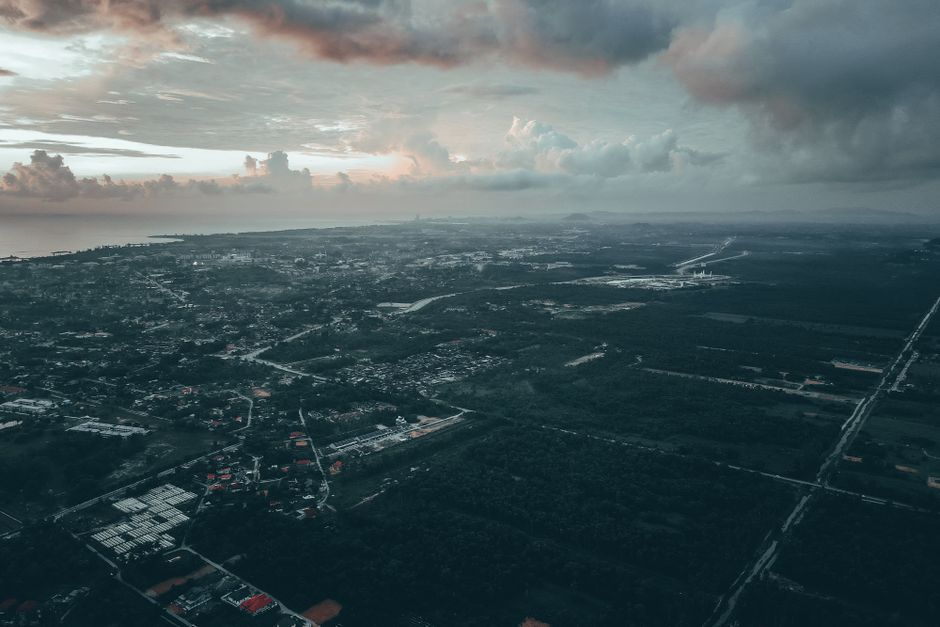 Breathtaking aerial view of city with residential buildings and lush green forests located near ocean under cloudy sunset sky