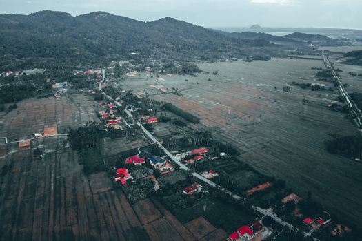 From above of small settlement located at roadside near agricultural fields surrounded by green mountains against cloudy sky