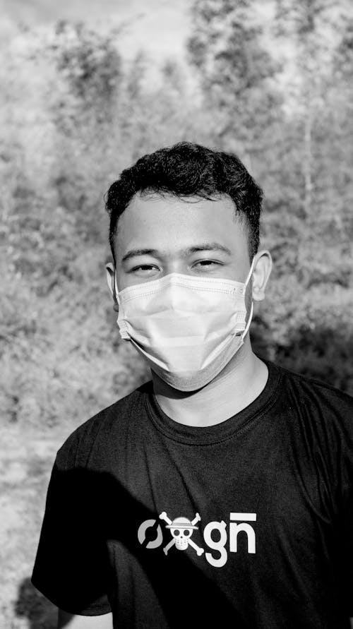 Bw of young Asian man wearing medical mask standing alone on green meadow in countryside and wearing casual clothes and looking at camera