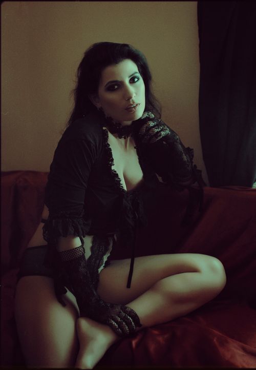 Free Seductive female with dark hair wearing seductive black bodysuit with gloves looking at camera while sitting on red couch in room Stock Photo