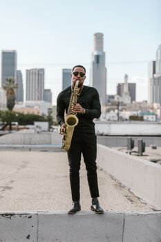 Black saxophonist playing on a rooftop with a city skyline background.