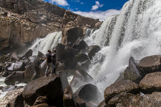 A couple standing on rocks by a cascading waterfall in Perú, enjoying nature's beauty.