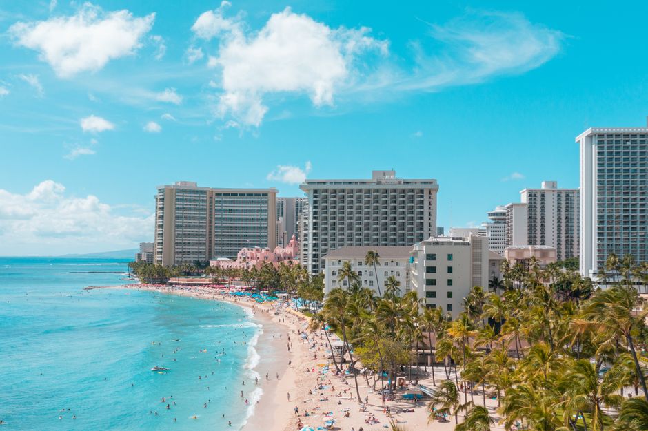 Photo by Jess Loiterton A vibrant aerial view of Waikiki Beach with luxurious hotels and sunlit turquoise ocean.