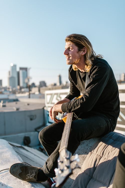 Free A young male musician sits on a rooftop, holding a guitar, overlooking the cityscape at sunset. Stock Photo