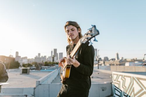 Free Young musician playing bass guitar on a rooftop with city skyline. Captivating urban ambiance. Stock Photo