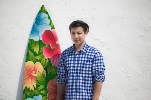 Man in Checkered Shirt Standing Beside a Surfboard 