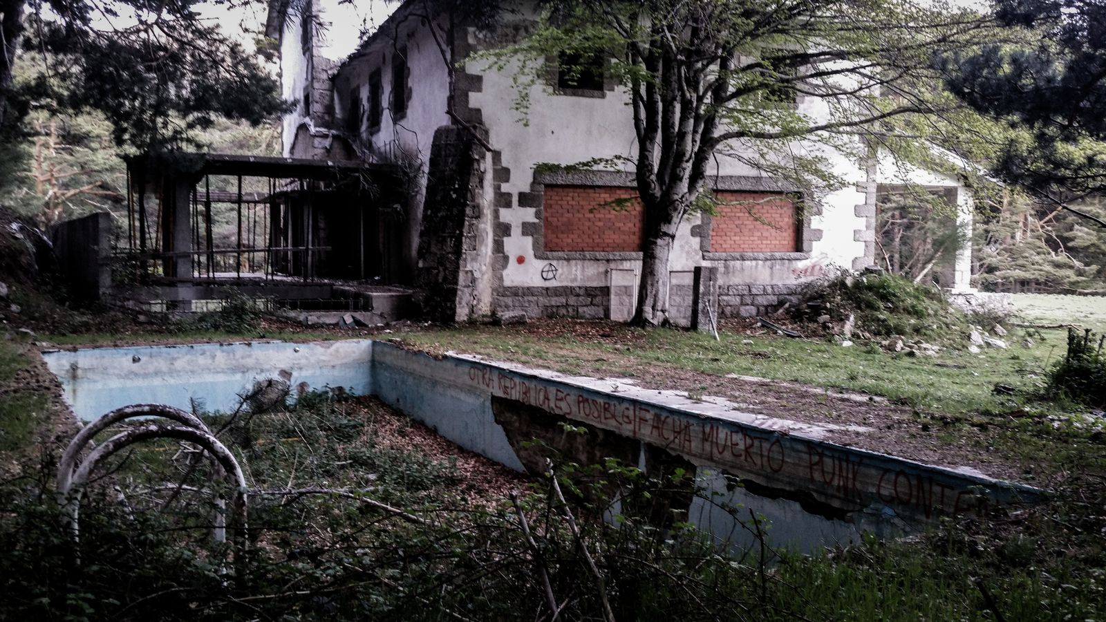 An overgrown, drained pool in front of a derelict building