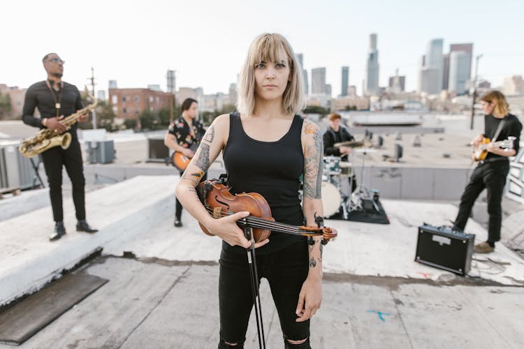 Woman In Black Top Holding Violin While Standing On The Rood With Music Band Behind