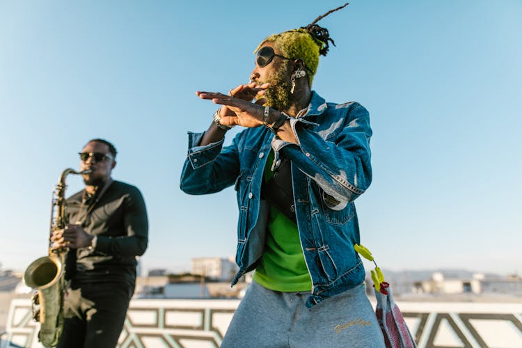 Man In Black Clothes Playing Saxophone Behind Man With Yellow Hair And Eyepatch Dancing