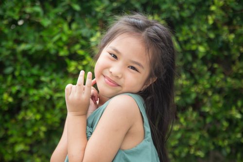 Free A cute Asian girl smiling and posing with a peace sign outdoors against a leafy background. Stock Photo
