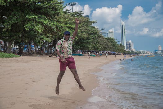 A man in a Hawaiian shirt jumps joyfully on a tropical beach with a cityscape in the background.