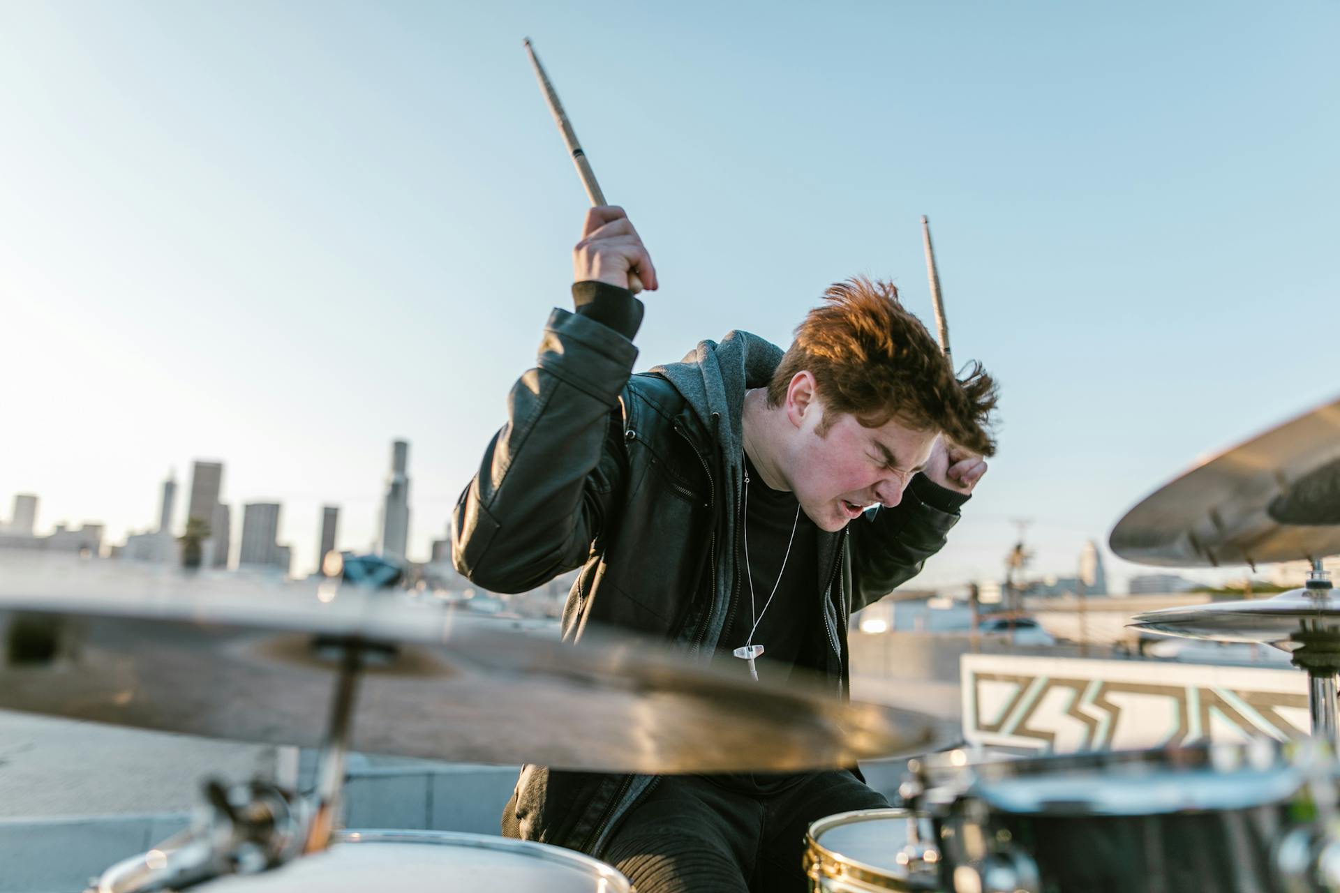 Musician in a black leather jacket passionately playing drums on a rooftop.