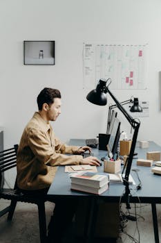 Man in brown jacket typing on a computer at a modern office desk with book stacks and lamps.