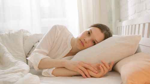 Free A woman in a white shirt lies on her side in bed, embracing a pillow, framed by soft natural lighting. Stock Photo