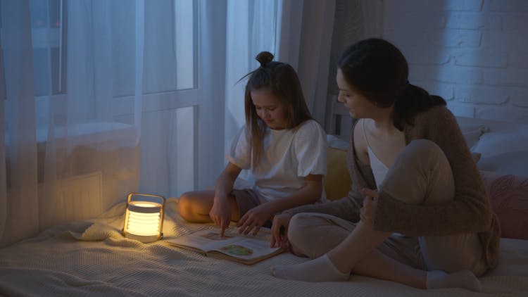 Daughter Sitting On Bed Beside Her Mother Reading A Book With A Night Lamp