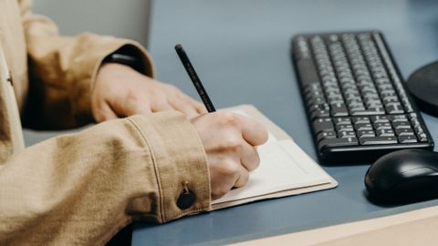 Close-up of hands writing in a notebook on an office desk with keyboard.