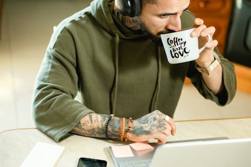 A Bearded Man in Green Hoodie Drinking Coffee from a Mug with Slogan