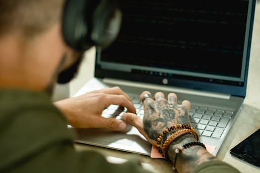 Tattooed freelancer working on a laptop, focused on coding tasks indoors. Modern tech lifestyle scene.