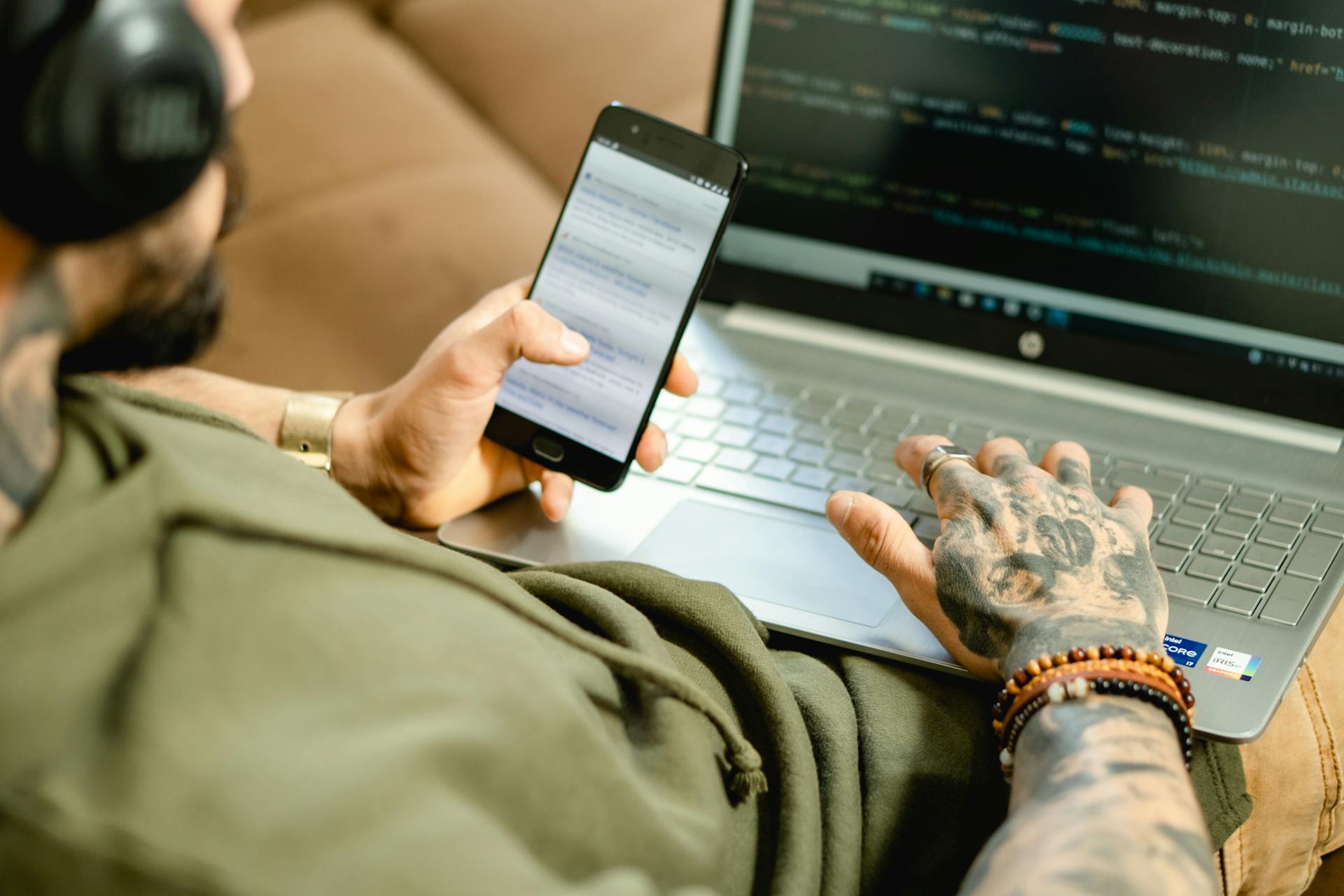 A tattooed person engaged in coding on a laptop while checking a smartphone.