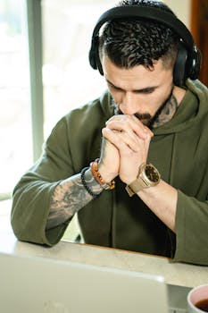 A focused man wearing headphones working on a laptop in a home office setting, deep in thought.
