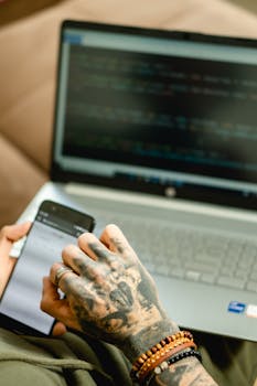 Close-up of a tattooed hand holding a smartphone, working on a laptop in an office setting.