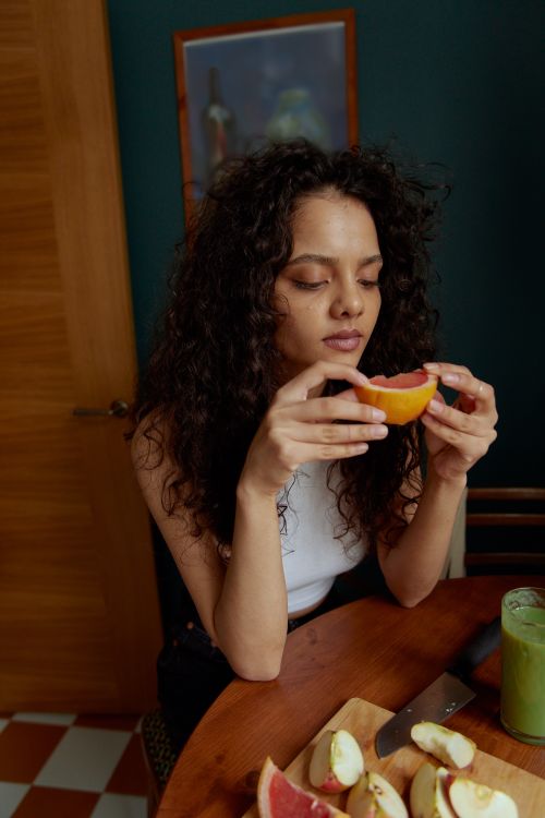 Free Young woman with curly hair holding a slice of citrus fruit at a table indoors. Stock Photo
