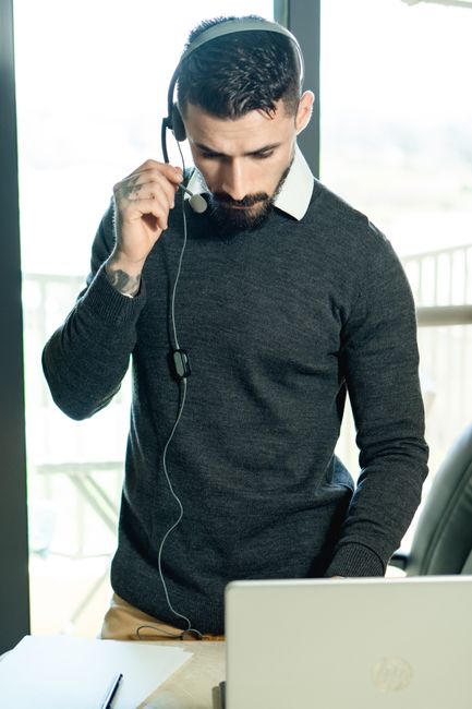 A bearded man in a home office setting uses a headset and laptop, conveying modern work tactics. A bearded man in a home office setting uses a headset and laptop, conveying modern work tactics.