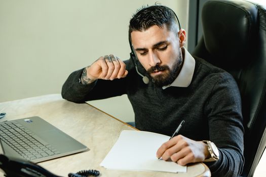 Man with beard and headset working diligently at desk with laptop and paper.