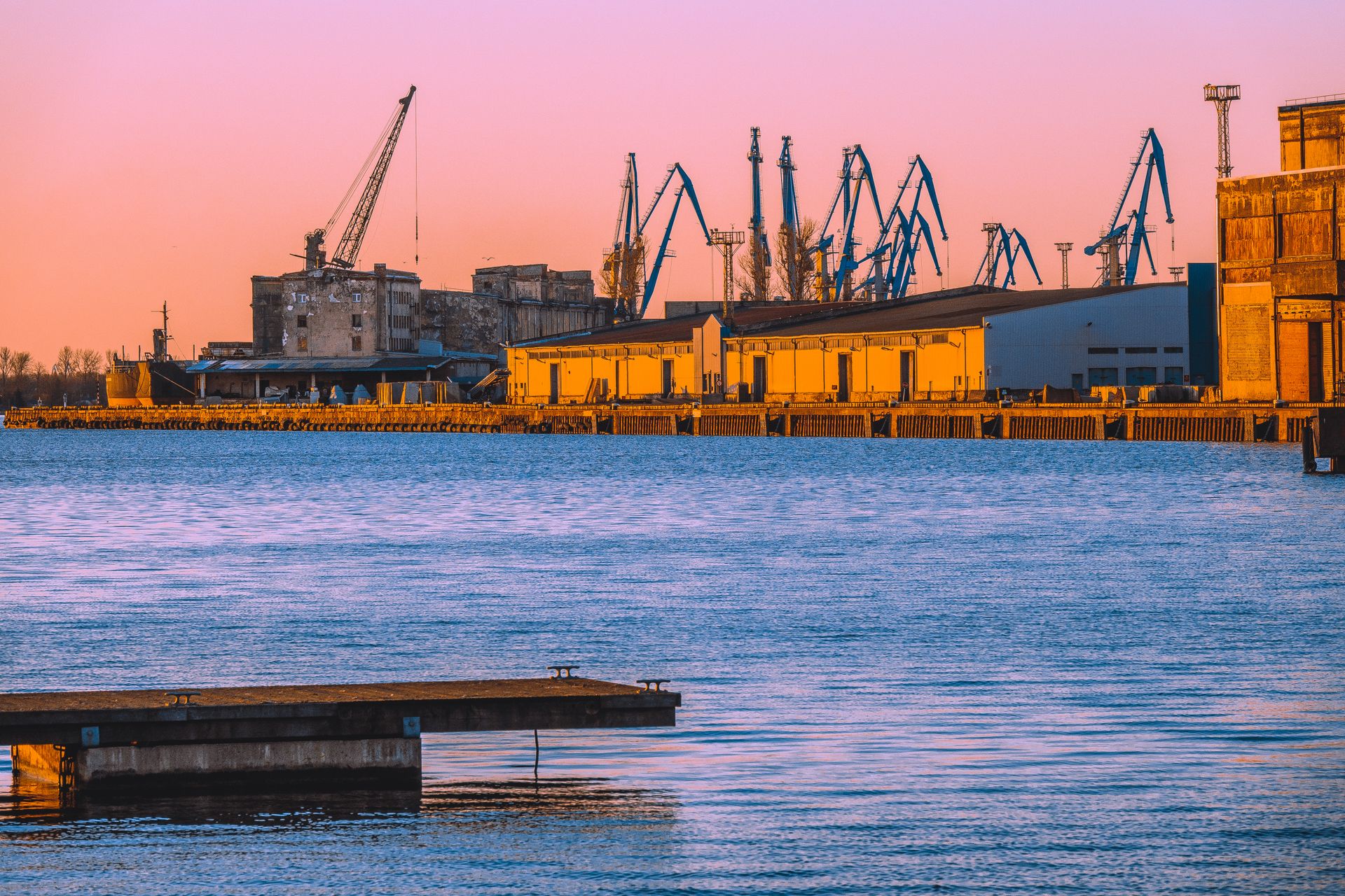 A vibrant industrial port scene in Riga, Latvia, with cranes silhouetted against a sunset sky.