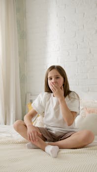Caucasian girl sitting on bed yawning, suggesting a sleepy morning or bedtime routine.