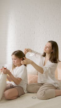 A mother gently combs her daughter's hair on a bed, capturing a tender family moment.