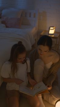 A mother and daughter share a warm moment reading a storybook by lamplight in a cozy bedroom setting.