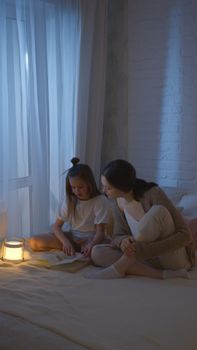 A mother and daughter share a quiet moment reading a book in a softly lit bedroom at night.